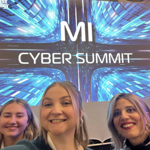 Cybersecurity students, Audry Douglas and Sophi Ernst, along with Dr. Samah Mansour smile for a selfie at the MI Cyber Summit, with a digital-themed event backdrop displaying "MI Cyber Summit" in bold text.